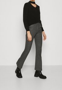 Kobieta w czarnym swetrze z wycięciami, szarych spodniach w jodełkę i czarnych botkach do kostki, stojąca na jasnym tle.