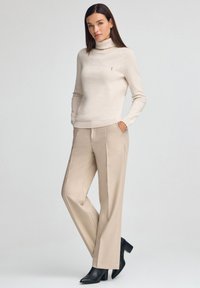 Beige coltrui gecombineerd met beige wijde broekspijpen. Slim fit, zachte textuur en subtiel logodetail. Zwarte enkellaarsjes met hak.