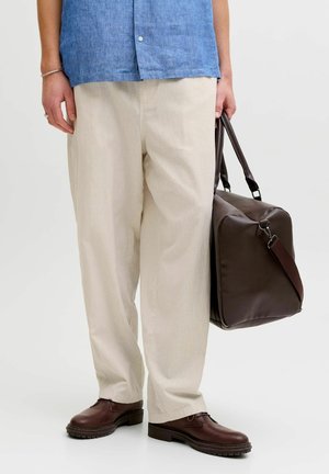 Egy személy bézs nadrágban, kék ingben és barna cipőben nagy sötétbarna bőr táskát tart a fogantyúinál fogva.