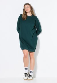 Eine Frau, die ein dunkelgrünes, übergroßes Sweatshirtkleid, weiße Socken mit schwarzem Text und weiße Sneaker trägt, steht vor einer weißen Wand.
