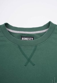 Grön sweatshirt med ribbad rundhals, svart etikett med texten "ÄRLIGHET" och texturerad sömnad över bröstet. Mjuk, blandning av bomullsmaterial.