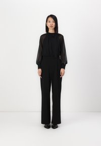 Zwart outfit bestaande uit een doorzichtige lange mouwen blouse met een gestrikte halslijn en hoge taille, wijde pantalon, aangevuld met platte zwarte schoenen.
