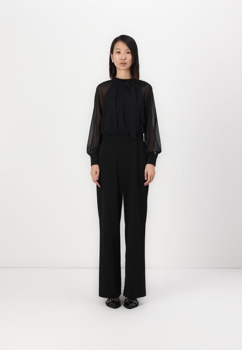 Zwart outfit bestaande uit een doorzichtige lange mouwen blouse met een gestrikte halslijn en hoge taille, wijde pantalon, aangevuld met platte zwarte schoenen.