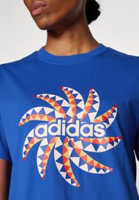 Blå bomullströja med grafiskt tryck som har ett flerfärgat geometriskt mönster och ordet "adidas" i vitt, rund halsringning.