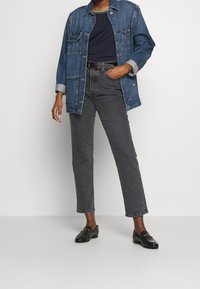 Jeansová bunda středně modré barvy s knoflíkovým zapínáním a dvěma náprsními kapsami, kombinovaná s tmavě šedými rovnými kalhotami a černými mokasíny.