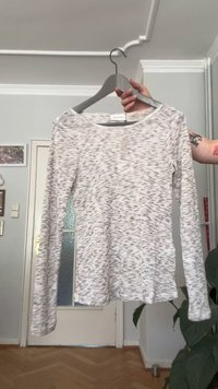 Light grey long-sleeve knit shirt with subtle darker speckles, held on a grey hanger in an indoor room with wooden floor and white walls.
