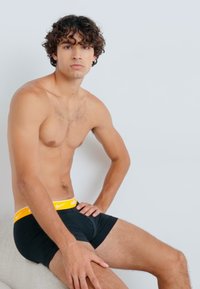 Boxers negros ajustados con una cinturilla amarilla de Nike, tela suave, ajuste ceñido, modelo sentado con una mano en la cadera, mirando hacia adelante.