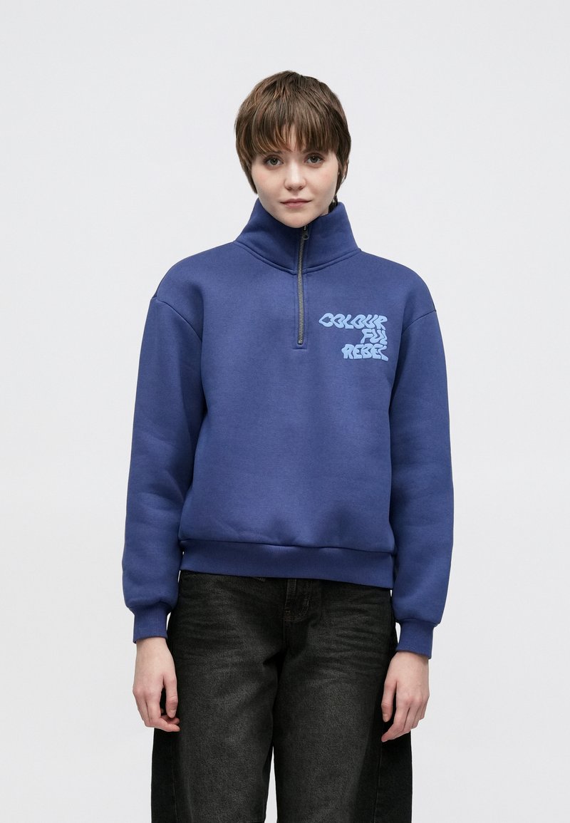 Zils fliisa džemperis ar augstu apkakli un pusējo rāvējslēdzēju, priekšpusē ar gaiši zilu tekstu "Colourful Rebels". Melnas džinsa bikses papildina izskatu.