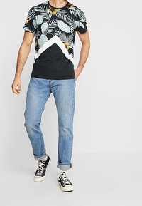 Muž v černém květinovém tričku, světle modrých vyhrnutých džínách a černých teniskách s bílými tkaničkami stojí na šedé podlaze.