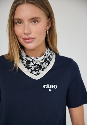 Junge Frau mit hellbraunen Haaren trägt goldene Creolen, ein schwarz-weiß gemustertes Halstuch und ein marineblaues Hemd mit „ciao“ und einem kleinen Herz.