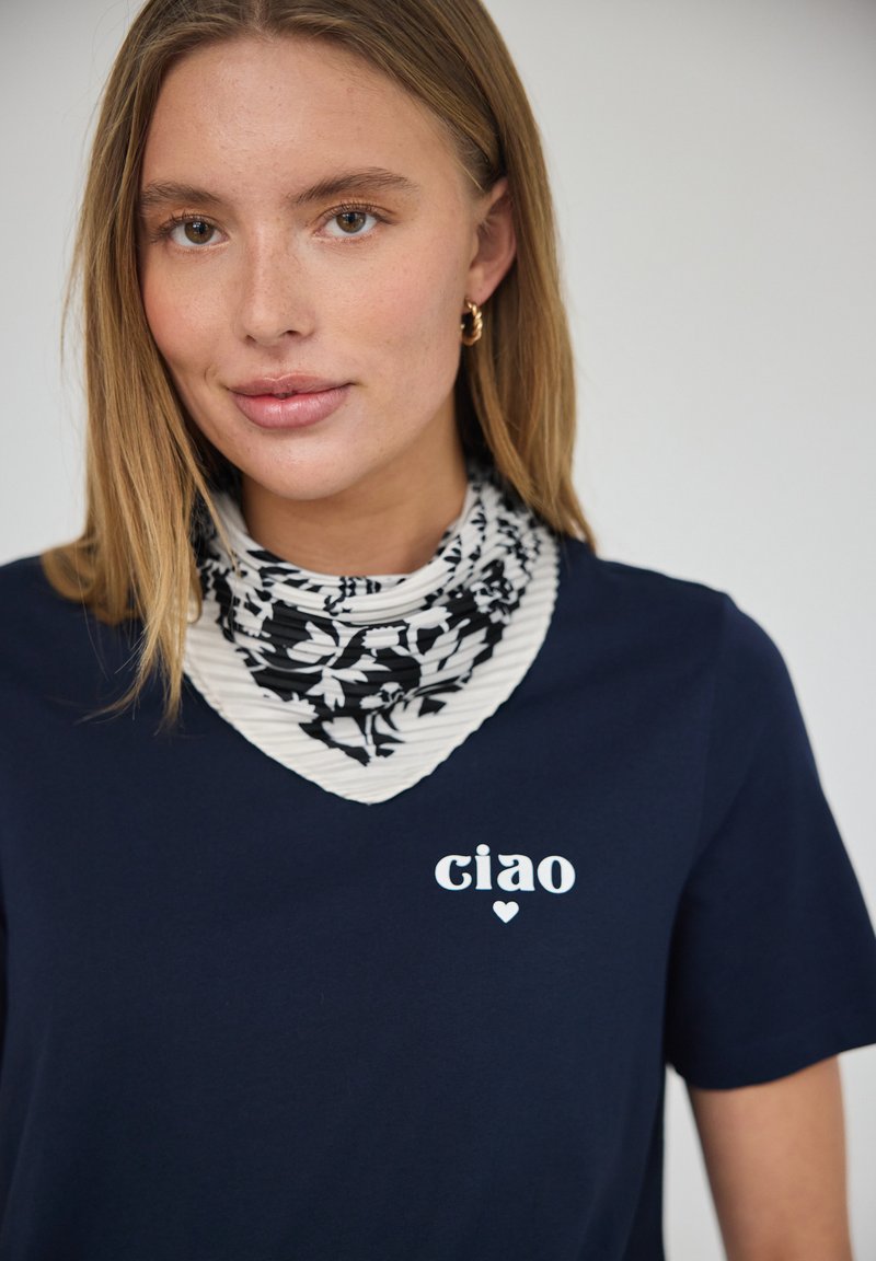 Jeune femme aux cheveux châtain clair porte des boucles d’oreilles anneaux dorés, un foulard à motifs noir et blanc autour du cou, et une chemise bleu marine avec « ciao » et un petit cœur.
