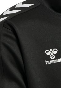 Îmbrăcăminte sport din material negru cu logo alb hummel pe piept și model alb în formă de chevron pe umăr.