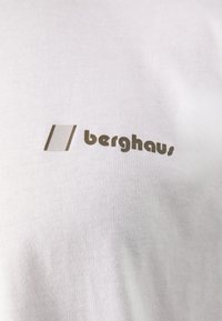 Fehér pamut póló, amelyen egy egyszerű barna logó található, ami "berghaus" feliratot tartalmaz, a szöveg felett pedig két függőleges vonal látható.