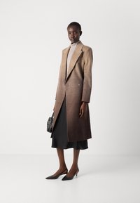 Lange, gradient bruine wollen jas met een notch-lapel en een dubbele knoopsluiting. Gecombineerd met een zwart satijnen slipdress en zwart gespte hakken.