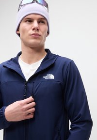 Νεαρός άνδρας με ανοιχτό μωβ σκουφάκι και γυαλιά ηλίου, που ρυθμίζει το φερμουάρ σε ένα μπλε σκούρο μπουφάν The North Face πάνω από λευκό πουκάμισο.