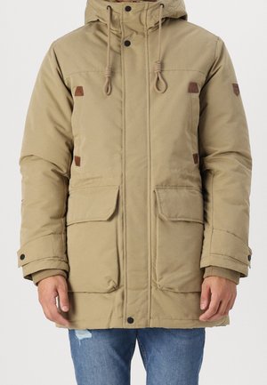 Beige vinterjakke med hætte, snore, trykknapper og to store forlommer, båret med blå jeans.