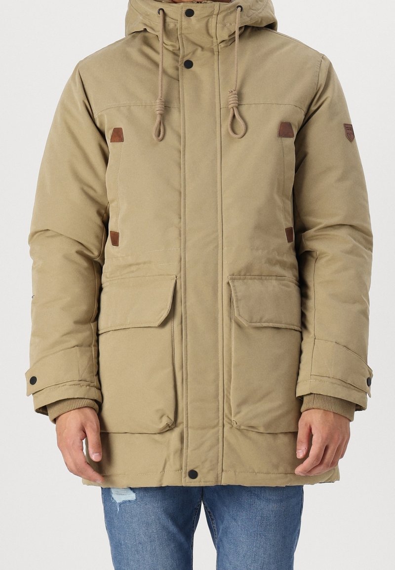 Veste d'hiver beige à capuche avec cordons, boutons-pression et deux grandes poches avant, portée avec un jean bleu.