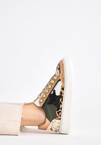Pie con una zapatilla multicolor con estampado animal, tachuelas, detalle de estrella negra y pantalones beiges sobre un fondo blanco.