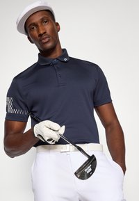 Marine polo skjorte med mønstrede ermer, hvit belte og lyse bukser. Holder en golfkølle, iført en hvit caps og golfhanske.