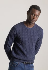 Suéter de punto trenzado azul marino con cuello redondo y puños acanalados, combinado con jeans de mezclilla azul, que muestran un patrón texturizado.