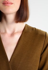 Bezruký top s hlubokým V- výstřihem v olivové barvě, vyrobený z texturované tkaniny, s krátkými rukávy a čistými švy.