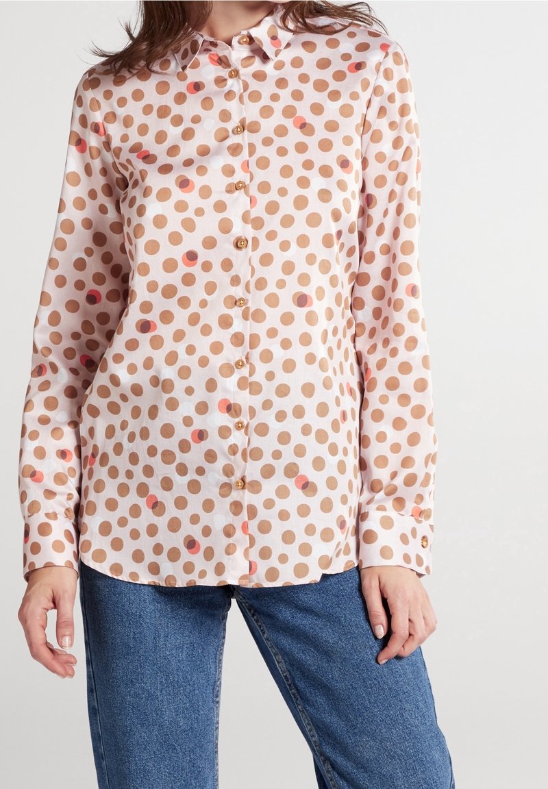 Femme portant un chemisier blanc à manches longues avec des pois marron et un jean bleu, debout devant un fond clair uni.