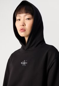 Čierna mikina s kapucňou vyrobená z jemného materiálu, s strieborným logom "Calvin Klein" a veľkou kapucňou. Jednoduchý dizajn bez viditeľných vzorov.