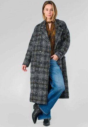 Long manteau croisé en motif pied-de-poule gris et noir, avec de grands revers et des poches latérales, porté sur un haut marron et un jeans bleu.