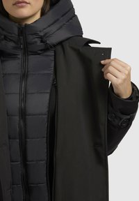 Čierna puffová bunda pod tmavým, jemne textúrovaným vonkajším kabátom. Predný zips s gombíkovými detailmi a kapucňa s elastickou šnúrkou.