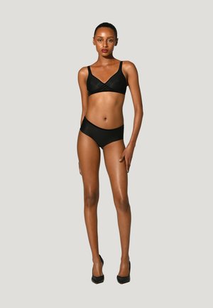 Donna in piedi, indossa reggiseno nero e biancheria nera, con la mano destra appoggiata sulla coscia, su sfondo grigio chiaro uniforme.