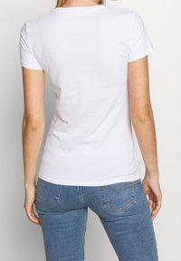 White cotton t-shirt with short sleeves, fitted design and crew neck. Back shows subtle logo at the neckline. Worn with blue jeans.