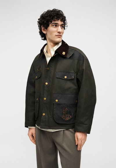 Άνδρας με σγουρά μαλλιά και γυαλιά, φοράει σκούρο πράσινο σακάκι με ορείχαλκα κουμπιά και κεντημένη μπροστινή τσέπη πάνω σε λευκό πουκάμισο.