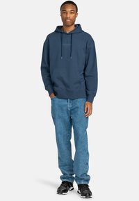 Navy blue hooded sweatshirt in soft fabric with a kangaroo pocket, paired with light denim jeans and black athletic shoes.