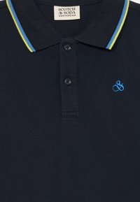 Mørk marineblå polo skjorte med klassisk krage, to-knaps placket og blå og gulestriper. Brodert logo på brystet.