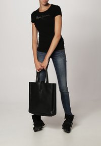 Nainen, joka pitää mustaa t-paitaa ja sinisiä farkkuja, pitelee molemmilla käsillään suurta mustaa nahkakassia tasaisella taustalla.