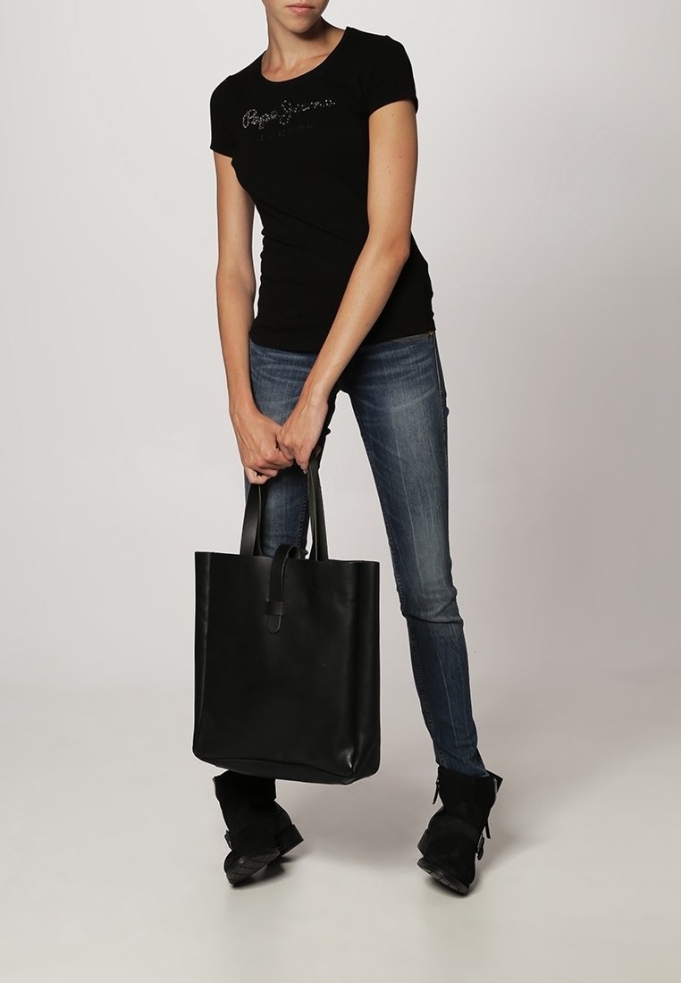 Nainen, joka pitää mustaa t-paitaa ja sinisiä farkkuja, pitelee molemmilla käsillään suurta mustaa nahkakassia tasaisella taustalla.