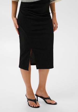 Femme portant une jupe midi noire avec une fente devant, un top gris clair et des sandales à talons noirs, debout sur un sol blanc.