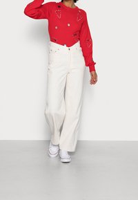 Rode trui met decoratieve patronen, witte wijde jeans en witte sneakers. De trui heeft geborduurde harten en ornamenten.