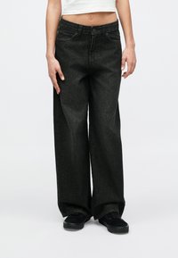 Άτομο που φοράει μαύρα τζιν με ψηλή μέση και άνετη εφαρμογή και μαύρα αθλητικά παπούτσια, στέκεται μπροστά σε απλό φωτεινό φόντο.