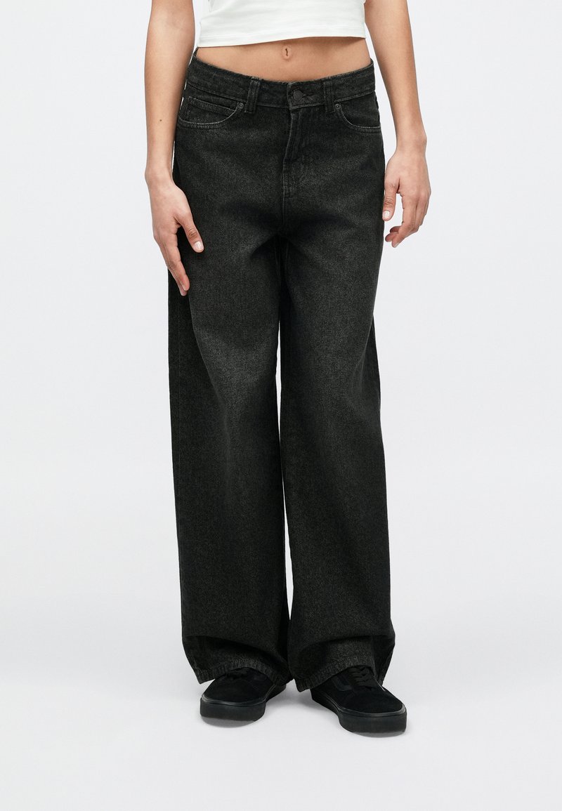 Person trägt hochgeschnittene, locker sitzende schwarze Jeans und schwarze Turnschuhe, steht vor einem schlichten hellen Hintergrund.