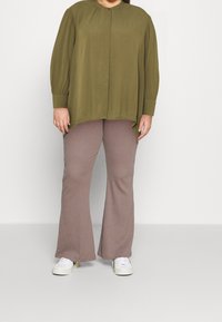 Blouse légère à manches longues de couleur vert olive, avec un col rond et une fermeture à boutons, associée à un pantalon évasé beige clair texturé et des baskets blanches.