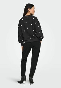 Sieviete redzama no mugurpuses, tērpusies melnā krekliņā ar baltām margrietiņu apdruku, melnās biksēs un melnās augstpapēžu potīšu kurpēs uz viendabīga fona.