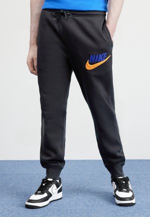 Osoba ubrana w czarne spodnie dresowe Nike z niebiesko-pomarańczowym logo, czarno-białe buty Nike, stojąca na niebieskim dywanie.
