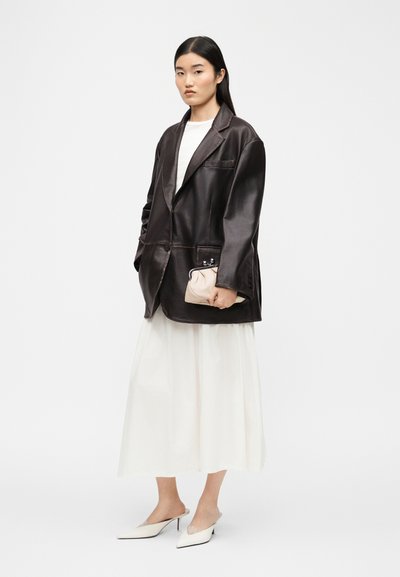 Γυναίκα που φοράει υπερμεγέθη σκούρο καφέ δερμάτινο σακάκι, λευκή μακριά φούστα, λευκές μυτερές γόβες, κρατώντας μικρό μπεζ πορτοφόλι μπροστά σε μονόχρωμο φόντο.