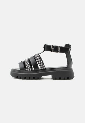 Sandale cu talpă groasă neagră, cu multiple bretele, detalii cu strasuri, cataramă la gleznă și închidere cu fermoar în spate, afișată pe un fundal alb.