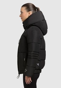 Sort oppustelig jakke med høj krave, quiltet design, frontlynlås og snørebånd i kanten. Har kontrastmærker på ærmerne.