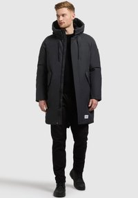 Černá vycpaná parka s kapucí, se zipem a zacvakáváním, bočními kapsami a logem poblíž spodního lemu.