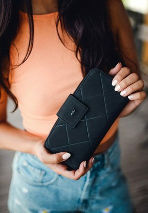 Black quilted wallet with a rectangular shape and a snap closure, held in a hand. Features a subtle embossed logo and smooth texture.