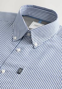 Camisa blanca a rayas azules, con cuello abotonado y etiqueta de marca. Presenta una sutil textura y múltiples botones en la parte frontal.