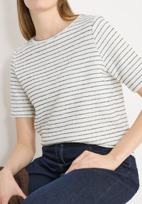 Femme portant un haut blanc à manches courtes avec de fines rayures horizontales noires, associé à un jean taille haute bleu foncé, assise sur un fond clair.